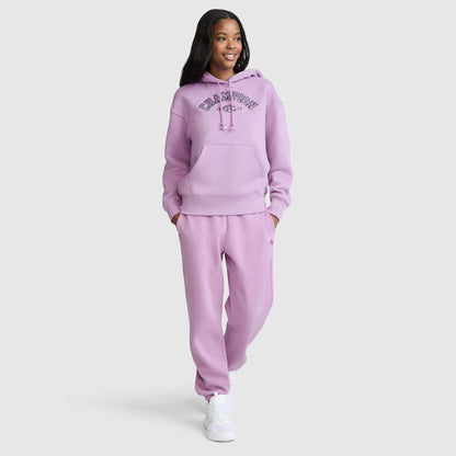 A person smiles while wearing a matching lavender Champion Fleece Hoodie, Satin Applique, and sweatpants set with white sneakers, standing against a plain white background.