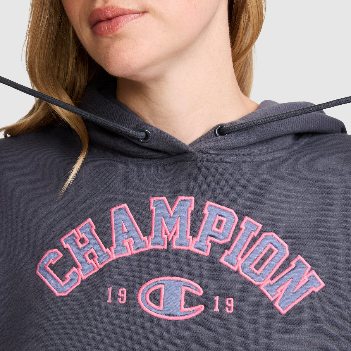 A woman is wearing a dark gray Champion Fleece Hoodie, Satin Applique, featuring "CHAMPION 1919" in pink outlined varsity-style letters on the front. Only her lower face and upper torso are visible.