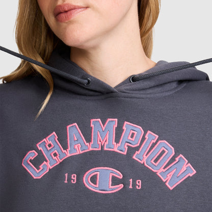 A woman is wearing a dark gray Champion Fleece Hoodie, Satin Applique, featuring "CHAMPION 1919" in pink outlined varsity-style letters on the front. Only her lower face and upper torso are visible.