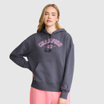A woman with long blonde hair wears a dark gray Champion Fleece Hoodie, Satin Applique, featuring pink varsity-style lettering and light pink sweatpants, standing against a plain light gray background.