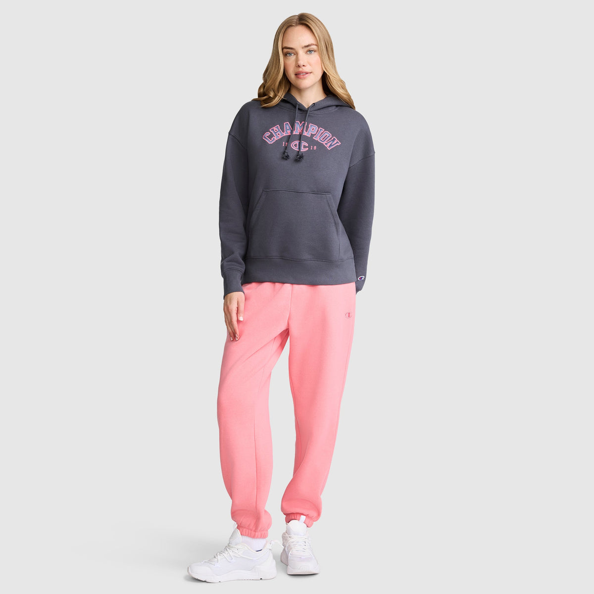 A woman stands against a plain gray background wearing a dark gray Champion Fleece Hoodie, Satin Applique, paired with light pink sweatpants and white sneakers. She has long, light brown hair and strikes a relaxed, casual pose.