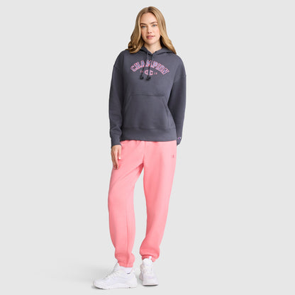 A woman stands against a plain gray background wearing a dark gray Champion Fleece Hoodie, Satin Applique, paired with light pink sweatpants and white sneakers. She has long, light brown hair and strikes a relaxed, casual pose.