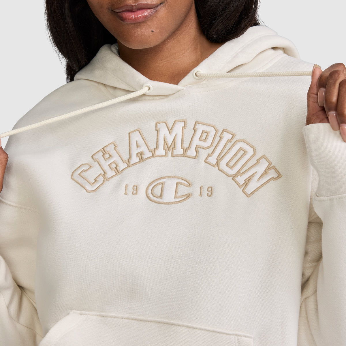 A woman wearing a cream-colored Champion Fleece Hoodie, Satin Applique, featuring "CHAMPION 1919" embroidered on the front in classic varsity style, gently holds the hoodie strings.