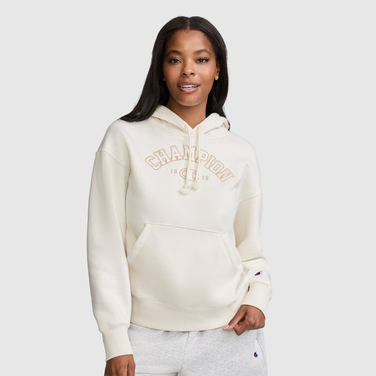 A woman stands against a plain background, smiling with one hand in her pocket, wearing a cream-colored Champion Fleece Hoodie with satin applique "CHAMPION" lettering on the front.