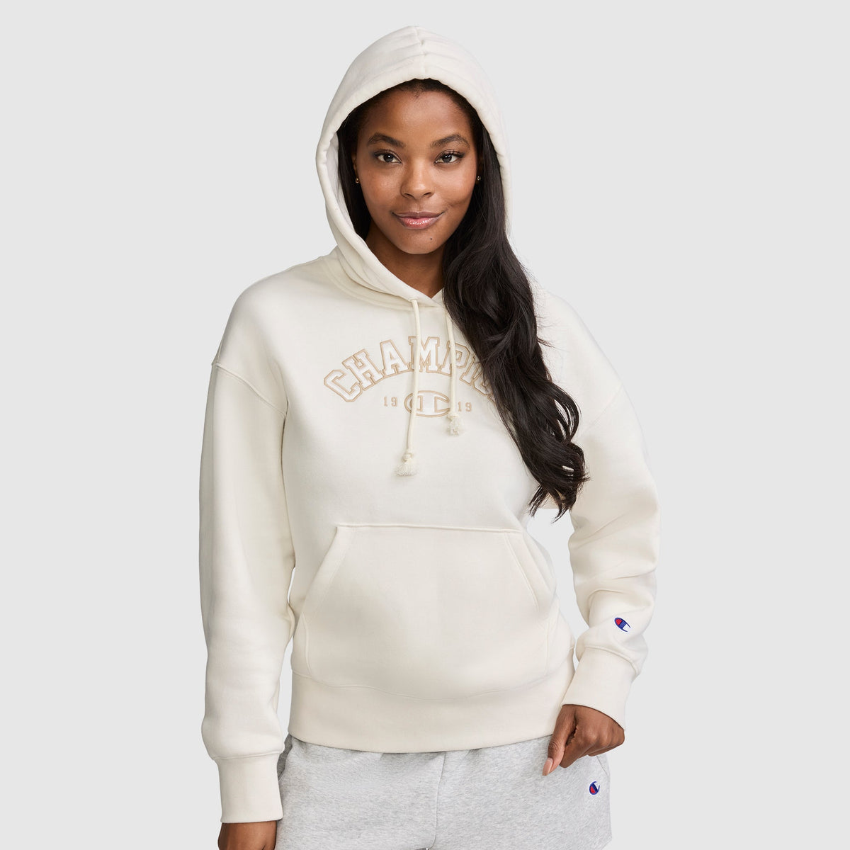 A woman with long dark hair and a relaxed expression stands against a plain light gray background, wearing the Champion Fleece Hoodie, Satin Applique in cream with the hood up. One hand is in her pocket as she showcases the varsity style hoodie featuring classic satin applique detailing for an elevated casual look.