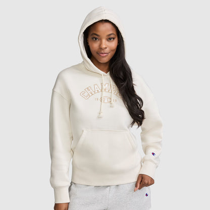 A woman with long dark hair and a relaxed expression stands against a plain light gray background, wearing the Champion Fleece Hoodie, Satin Applique in cream with the hood up. One hand is in her pocket as she showcases the varsity style hoodie featuring classic satin applique detailing for an elevated casual look.