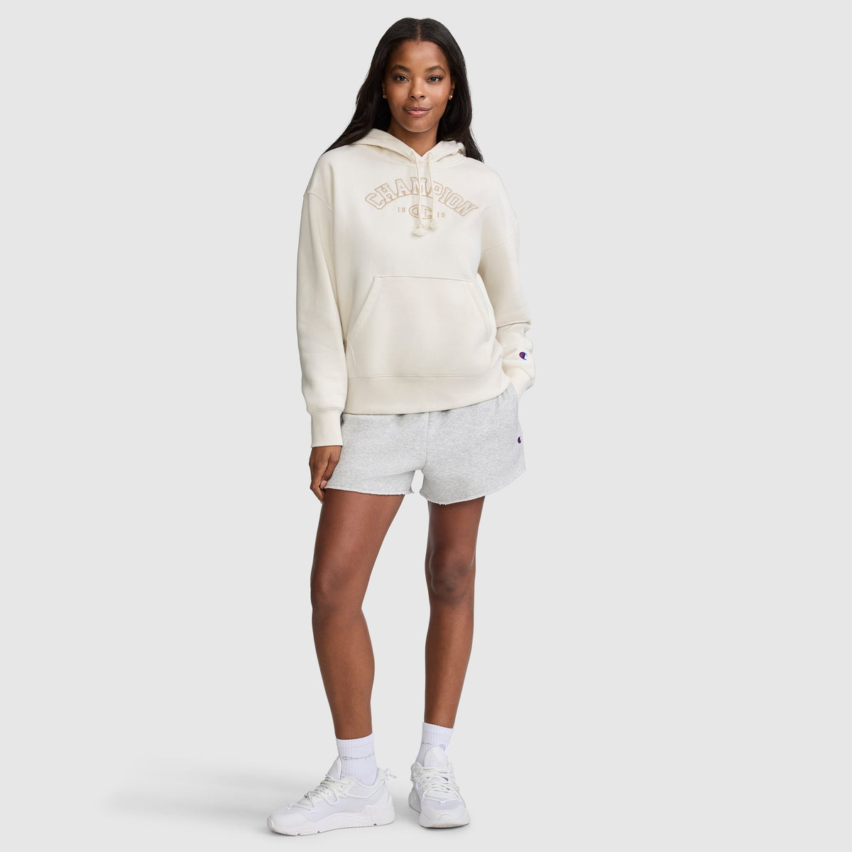 A woman stands facing forward with a neutral expression against a plain light background, wearing a cream Champion Fleece Hoodie, Satin Applique, paired with light gray shorts, white socks, and white sneakers.