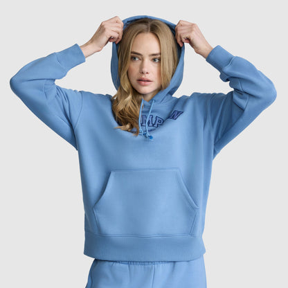 A woman wears the Champion Fleece Hoodie with Satin Applique and matching sweatpants, pulling up her hood as she faces forward against a plain background, with one hand on each side of the hood.