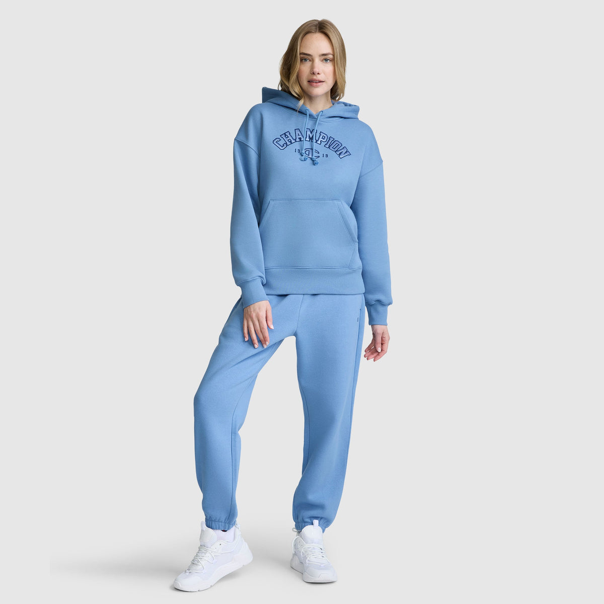 A person is wearing a light blue Champion Fleece Hoodie, Satin Applique, paired with matching joggers and white sneakers, standing against a plain light background.