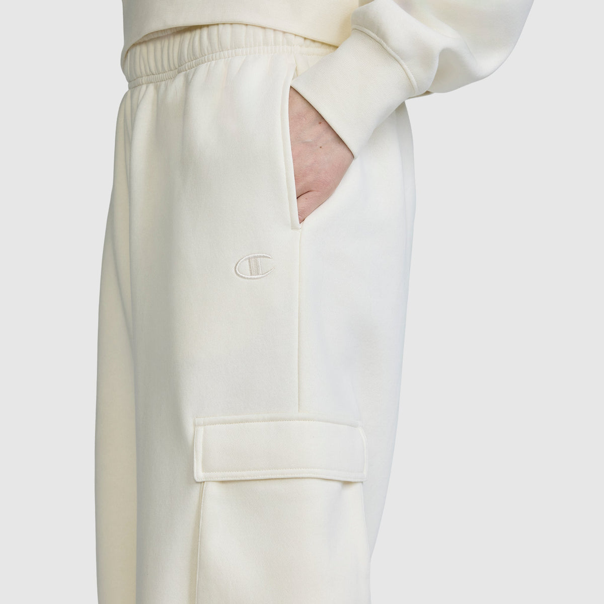 A person wearing the Champion Fleece Cargo Training Pant in off-white with a hand in the pocket. These pants showcase an embroidered C logo and a utility pocket with a flap on the thigh, combining style and functionality.