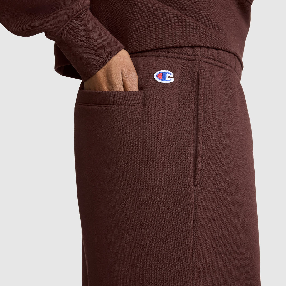 A person wearing brown Champion Fleece Cargo Training Pants and a matching sweatshirt has their hand in their pocket. The Champion logo is visible near the waistband, emphasizing the cozy style of the Fleece Cargo Training Pant by Champion.