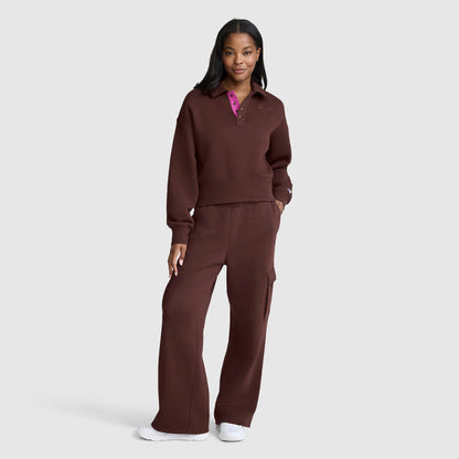 A woman stands smiling, wearing a matching dark brown pullover and Champion Fleece Cargo Training Pant, paired with white sneakers against a plain light gray background.