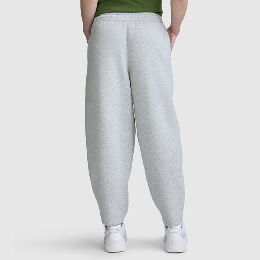 A person is wearing the Champion Barrel Leg Fleece Pant in light gray, paired with white sneakers and a green top. They are standing with hands behind their back, facing away from the camera against a plain background.