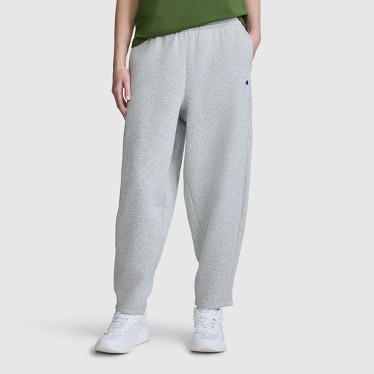 A person is shown from the waist down, wearing the Champion Barrel Leg Fleece Pant in light gray, paired with a green shirt and white sneakers, against a plain white background. The relaxed fit of these Champion women’s pants is prominently displayed.