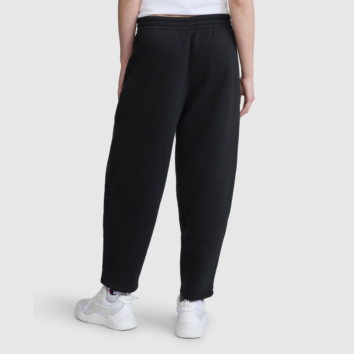 A person stands with their back to the camera against a plain light background, wearing Champion Barrel Leg Fleece Pants made from midweight cotton-poly fleece, paired with a white T-shirt and white sneakers.