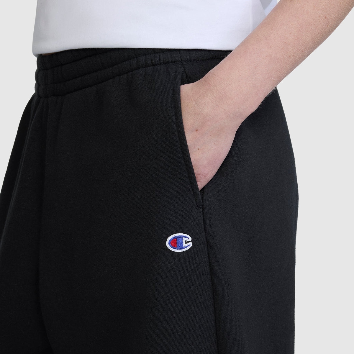 A person is seen wearing Champion's Barrel Leg Fleece Pant in black, with one hand tucked into the pocket. Only the lower torso and part of an arm are visible, showcasing the embroidered Champion logo on these midweight cotton-poly sweatpants.