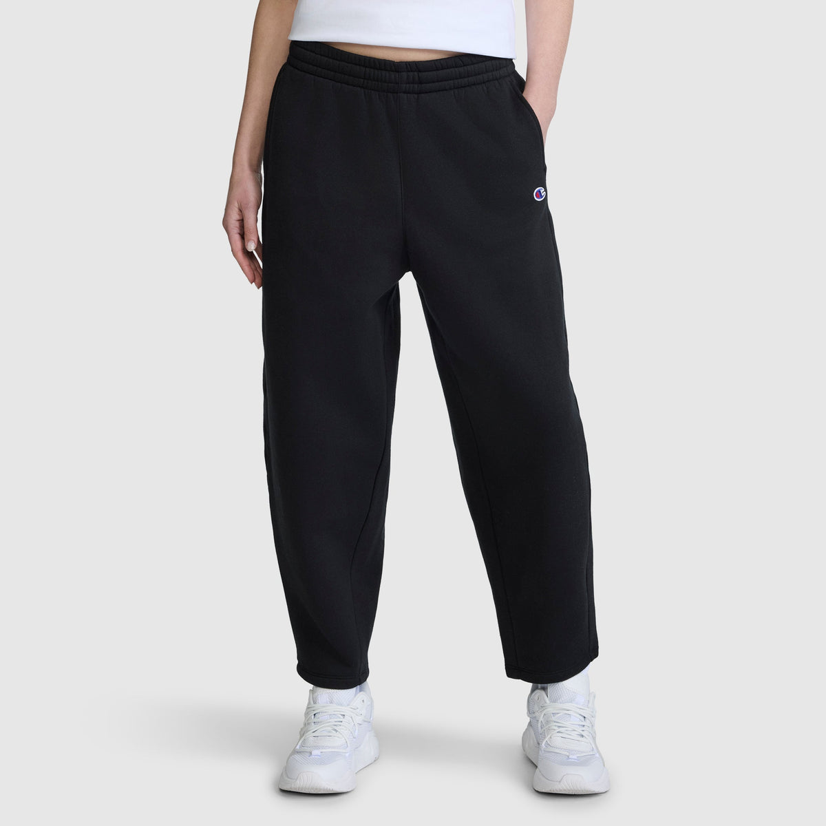 A person is wearing the Champion Barrel Leg Fleece Pant, crafted from midweight cotton-poly fleece in black, paired with a white cropped top and white sneakers, standing against a plain light gray background.
