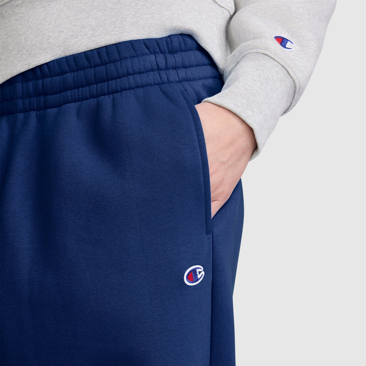 A person wearing a gray sweatshirt and blue Champion Barrel Leg Fleece Pants has their right hand in their pocket. The image highlights the waist and hip area, showcasing the midweight fleece fabric and the relaxed barrel leg style from Champion.