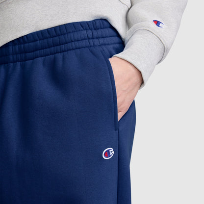 A person wearing a gray sweatshirt and blue Champion Barrel Leg Fleece Pants has their right hand in their pocket. The image highlights the waist and hip area, showcasing the midweight fleece fabric and the relaxed barrel leg style from Champion.