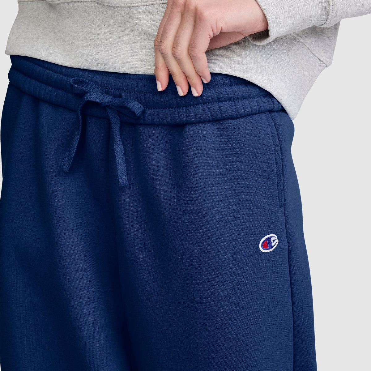 A person wearing a light gray sweatshirt and navy blue Champion Barrel Leg Fleece Pants adjusts the waistband with one hand. The midweight fleece offers cozy warmth, blending comfort and style in this casual Champion look.