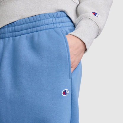 A person wearing a grey sweatshirt and blue Champion Barrel Leg Fleece Pants featuring contemporary sweats styling and subtle logos, with one hand in the pocket of the sweatpants.