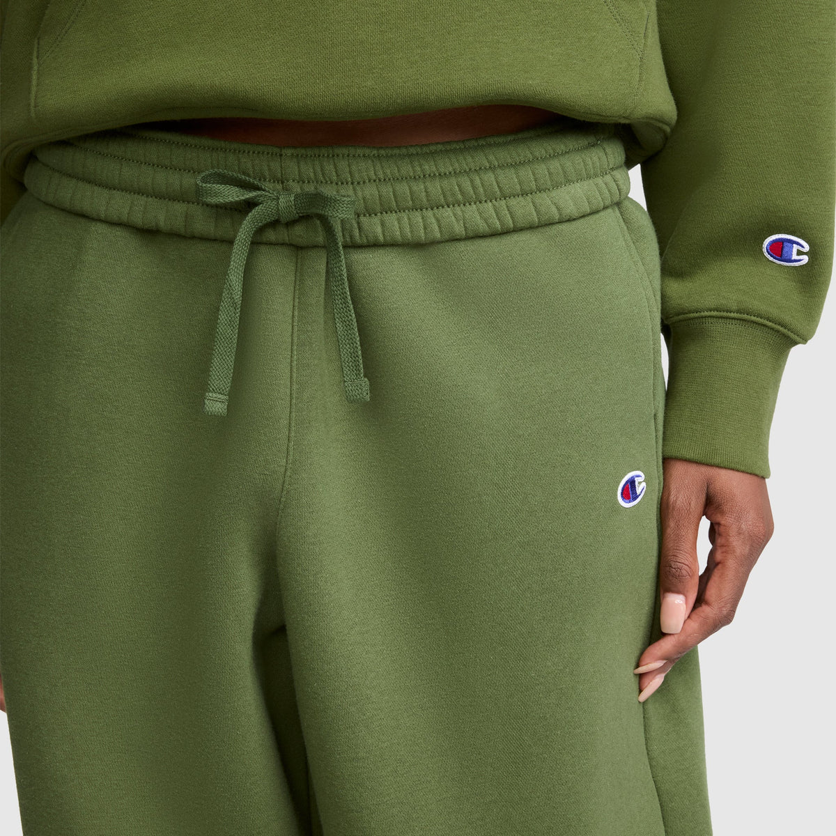 A person wears Champion Barrel Leg Fleece Pants in olive green along with a matching sweatshirt. One hand is visible by their side. Both garments display the Champion logo patches, and the midweight fleece fabric provides comfortable warmth.