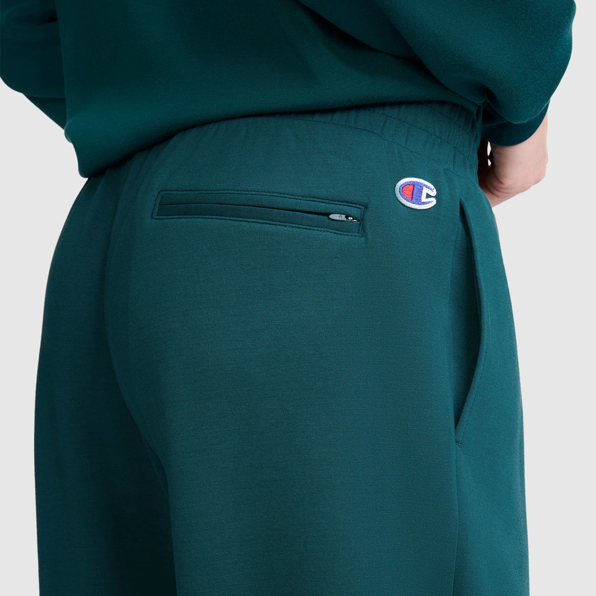 A close-up view shows a person wearing the Champion Taper Leg Jogger with Zippers, featuring a zippered back pocket and the signature Champion logo patch near the waistband. Their hand rests partially in the pocket, emphasizing the jogger’s comfortable fit and streamlined, tapered-leg design.