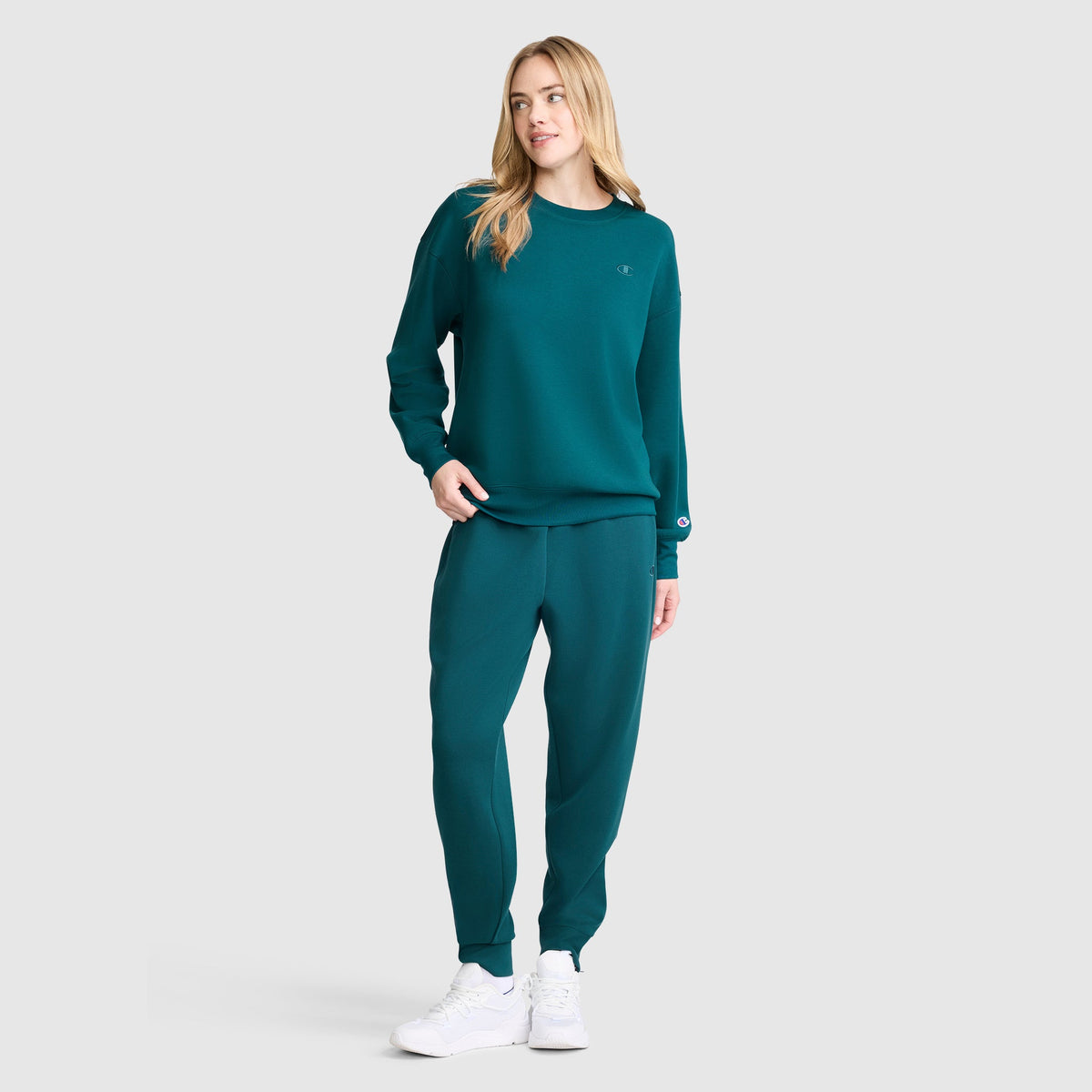 A woman stands wearing a matching teal Champion Taper Leg Jogger with Zippers and sweatshirt, paired with white sneakers, posing against a light gray background.