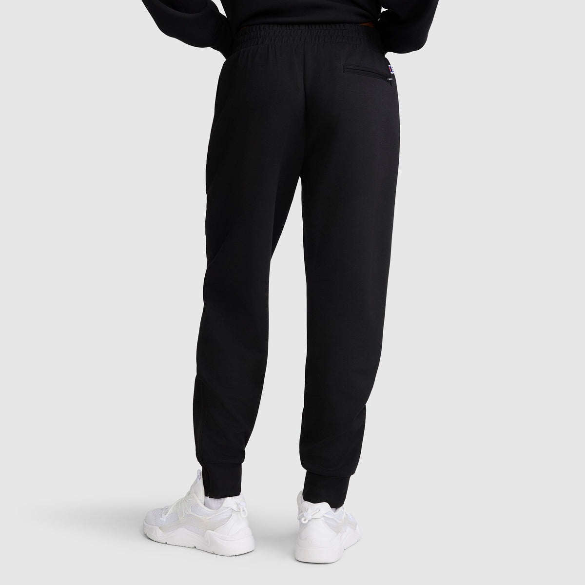 A person is seen from the back, standing against a plain light gray background, wearing the Champion Taper Leg Jogger with Zippers in black paired with white sneakers.