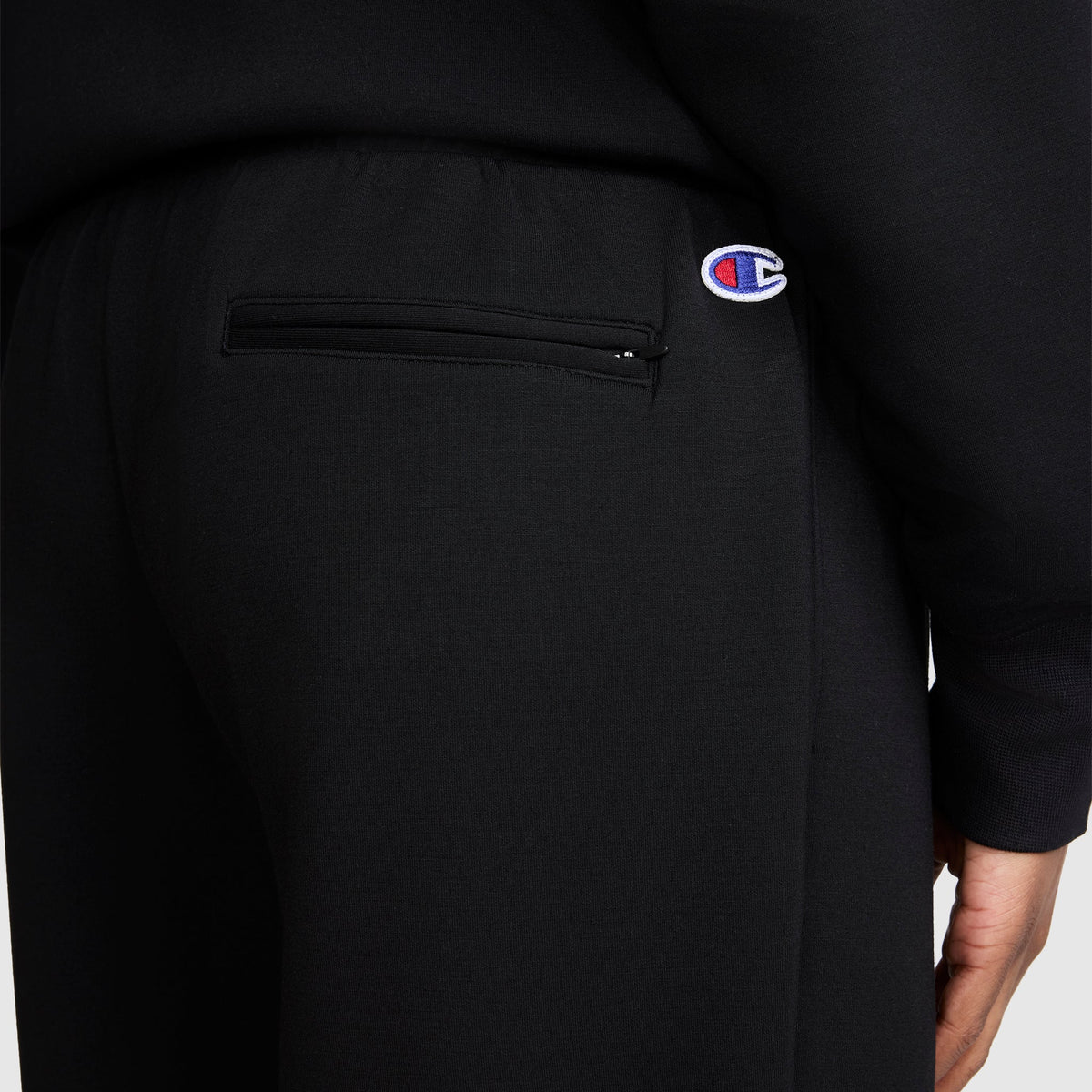 A close-up of a person wearing black Champion Taper Leg Jogger with Zippers, featuring a zippered back pocket and the Champion logo patch above it.