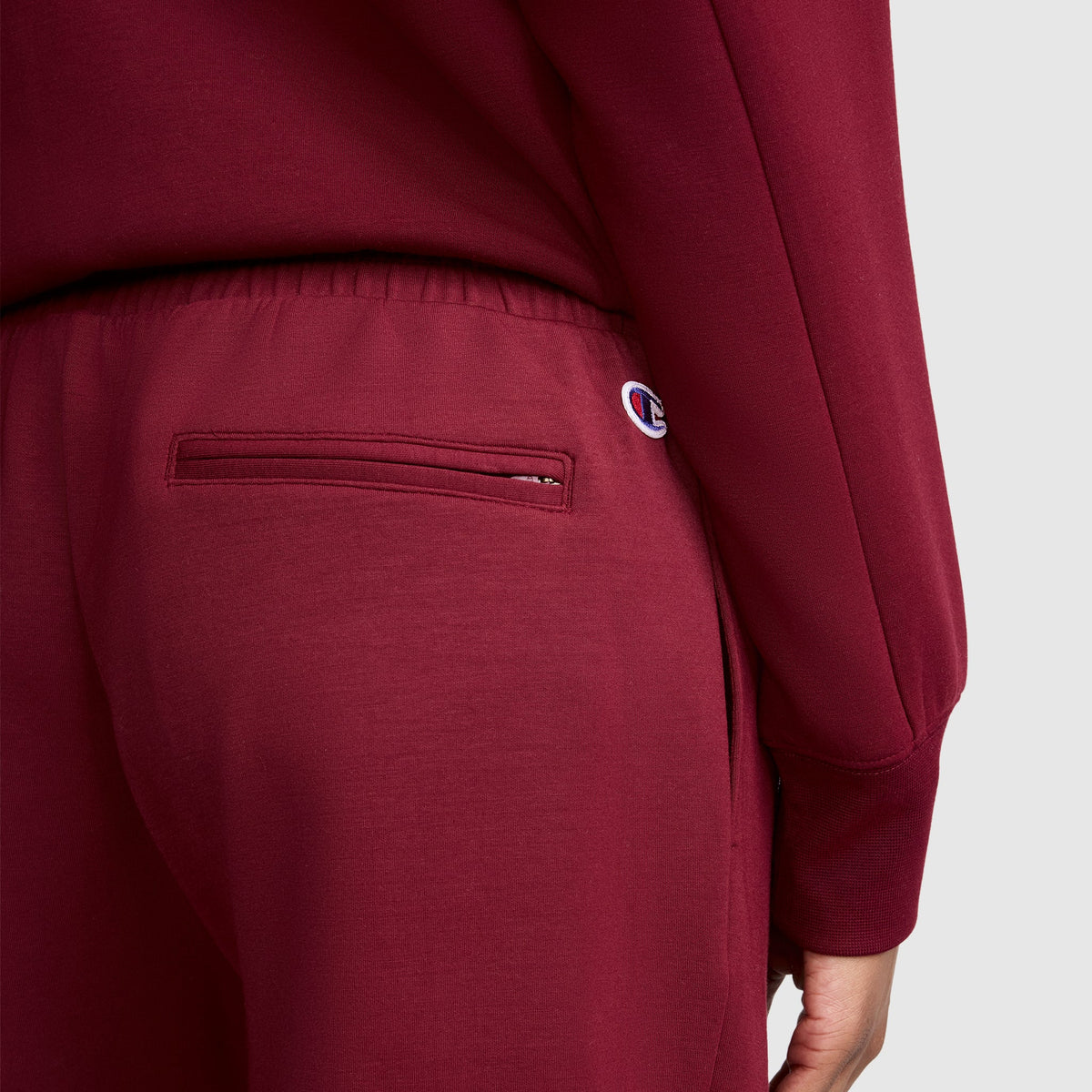 A person is wearing the Champion Taper Leg Jogger with Zippers in maroon, paired with a matching top. The joggers feature a small Champion logo patch near the right pocket, and their tapered leg design creates a flattering silhouette. Only the lower back and part of one arm are visible.