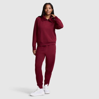 A woman stands against a plain background wearing a matching maroon Champion Taper Leg Jogger with Zippers set and white sneakers. She has one hand touching her hair and smiles slightly.