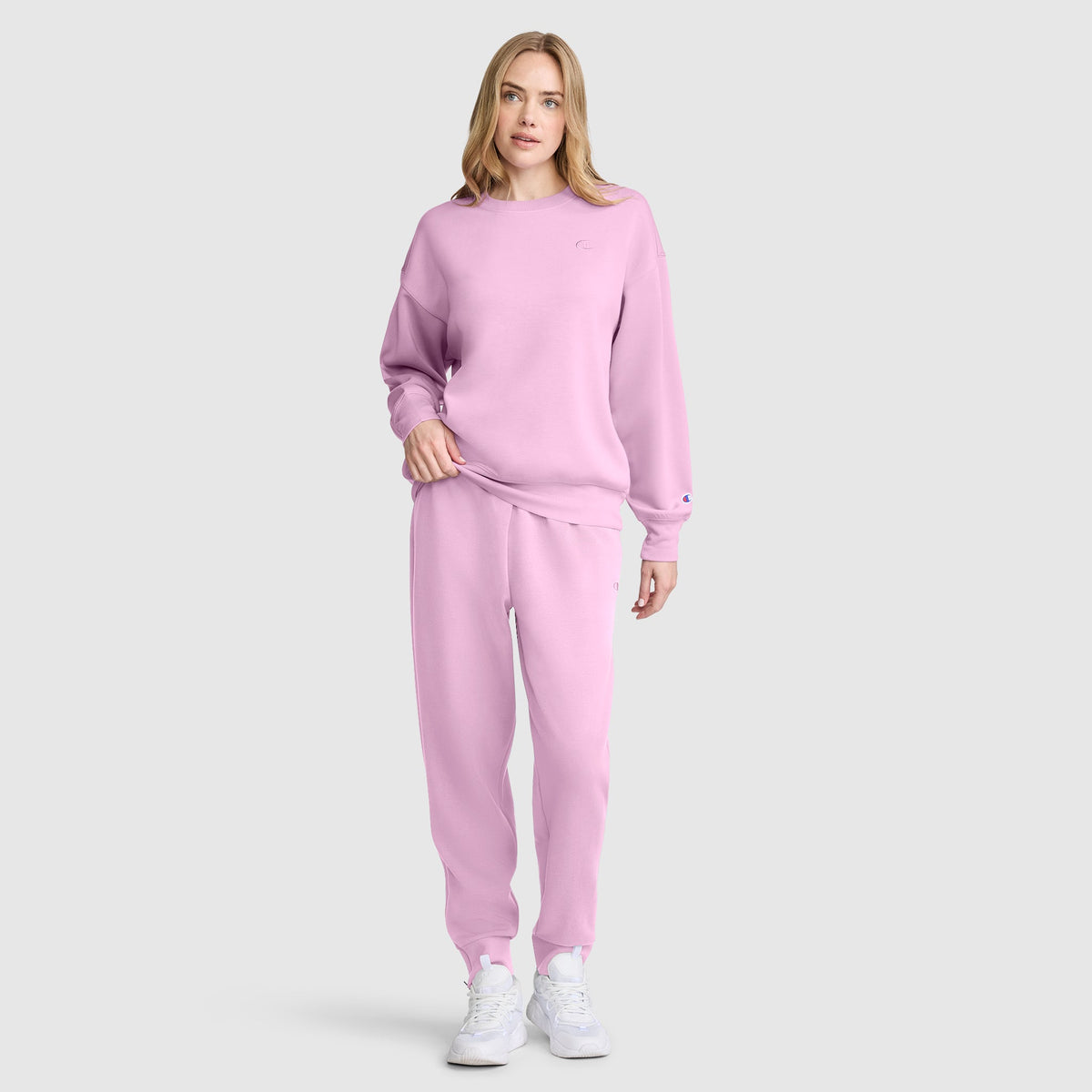 A woman stands against a plain gray background wearing a matching light pink Champion Taper Leg Jogger with Zippers set and white sneakers.