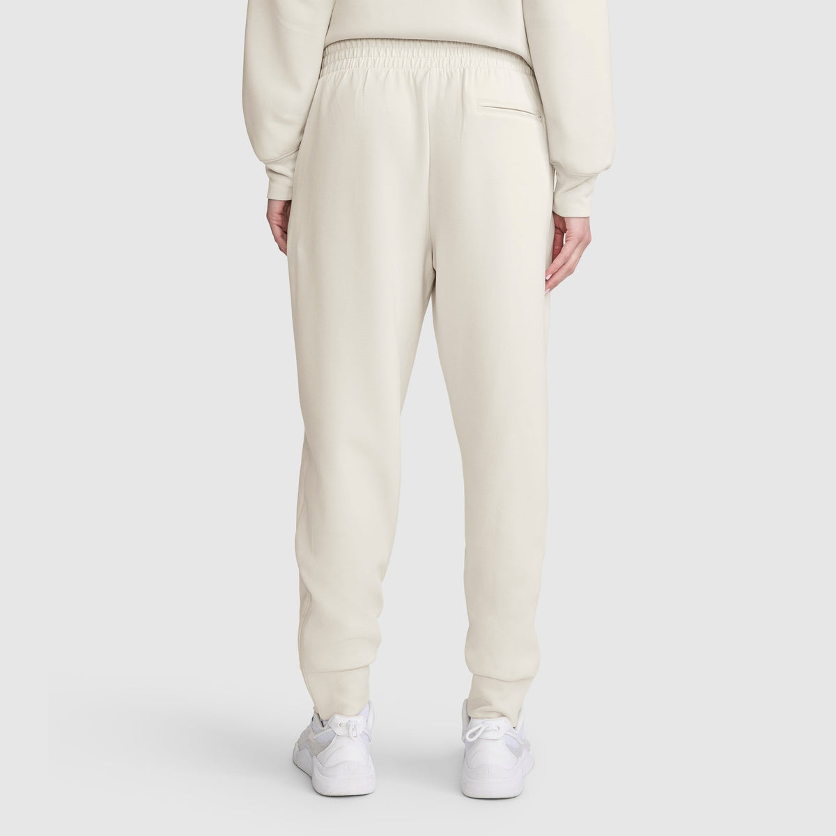 A person is seen from behind wearing cream-colored Champion Taper Leg Jogger with Zippers and a matching sweatshirt. The joggers feature a tapered leg, are made from a soft modal scuba blend, and have an elastic waistband. White sneakers complete the outfit, set against a plain light gray background.