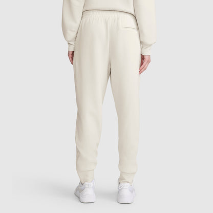 A person is seen from behind wearing cream-colored Champion Taper Leg Jogger with Zippers and a matching sweatshirt. The joggers feature a tapered leg, are made from a soft modal scuba blend, and have an elastic waistband. White sneakers complete the outfit, set against a plain light gray background.