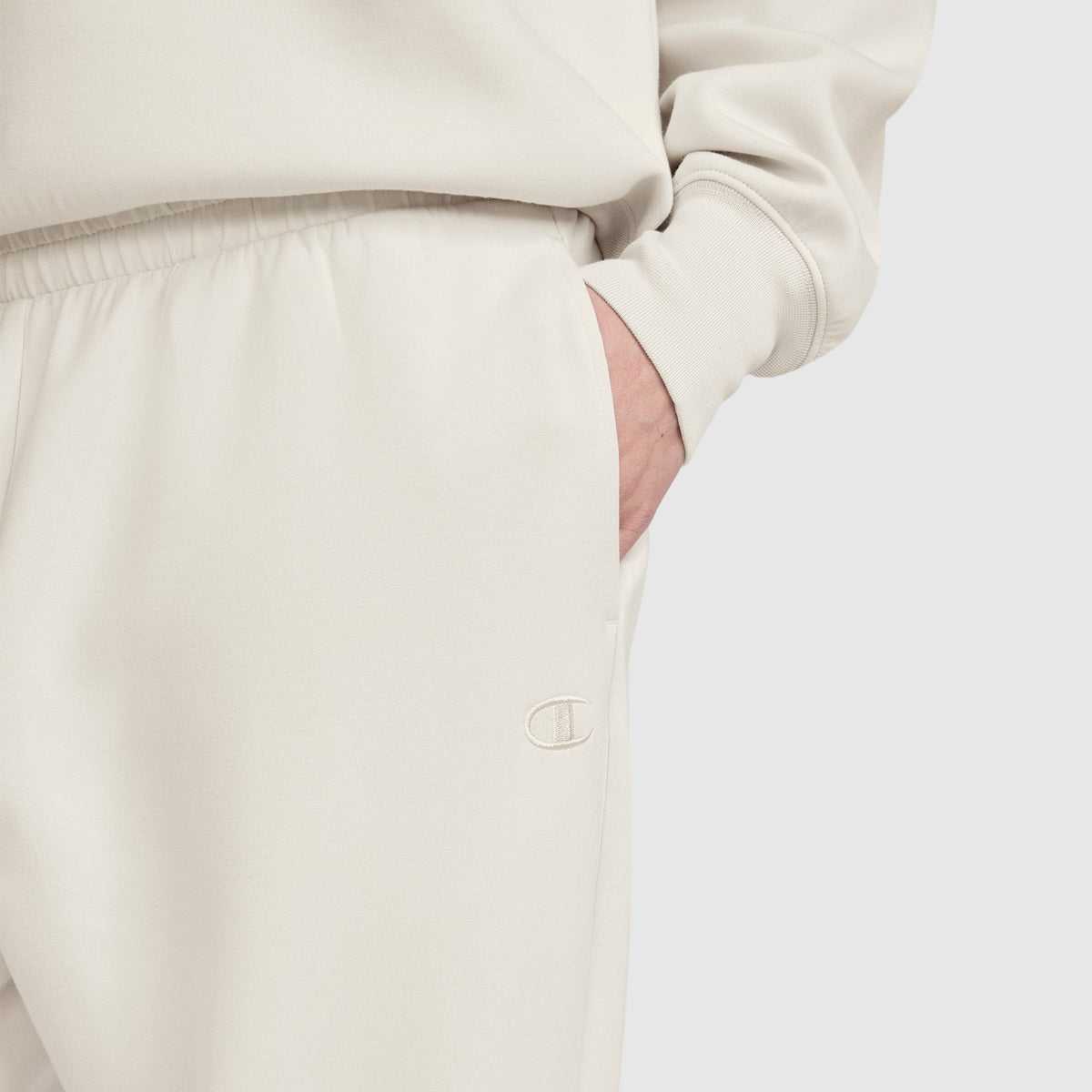 A person is wearing off-white Champion Taper Leg Jogger with Zippers and a matching sweatshirt, with one hand in the pocket. The Champion logo is embroidered on the jogger near the pocket, highlighting the signature style of Champion. The background is plain and light-colored.