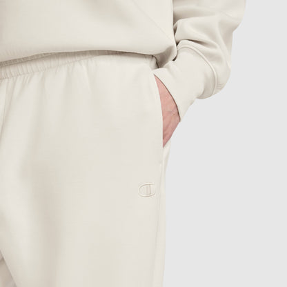 A person is wearing off-white Champion Taper Leg Jogger with Zippers and a matching sweatshirt, with one hand in the pocket. The Champion logo is embroidered on the jogger near the pocket, highlighting the signature style of Champion. The background is plain and light-colored.