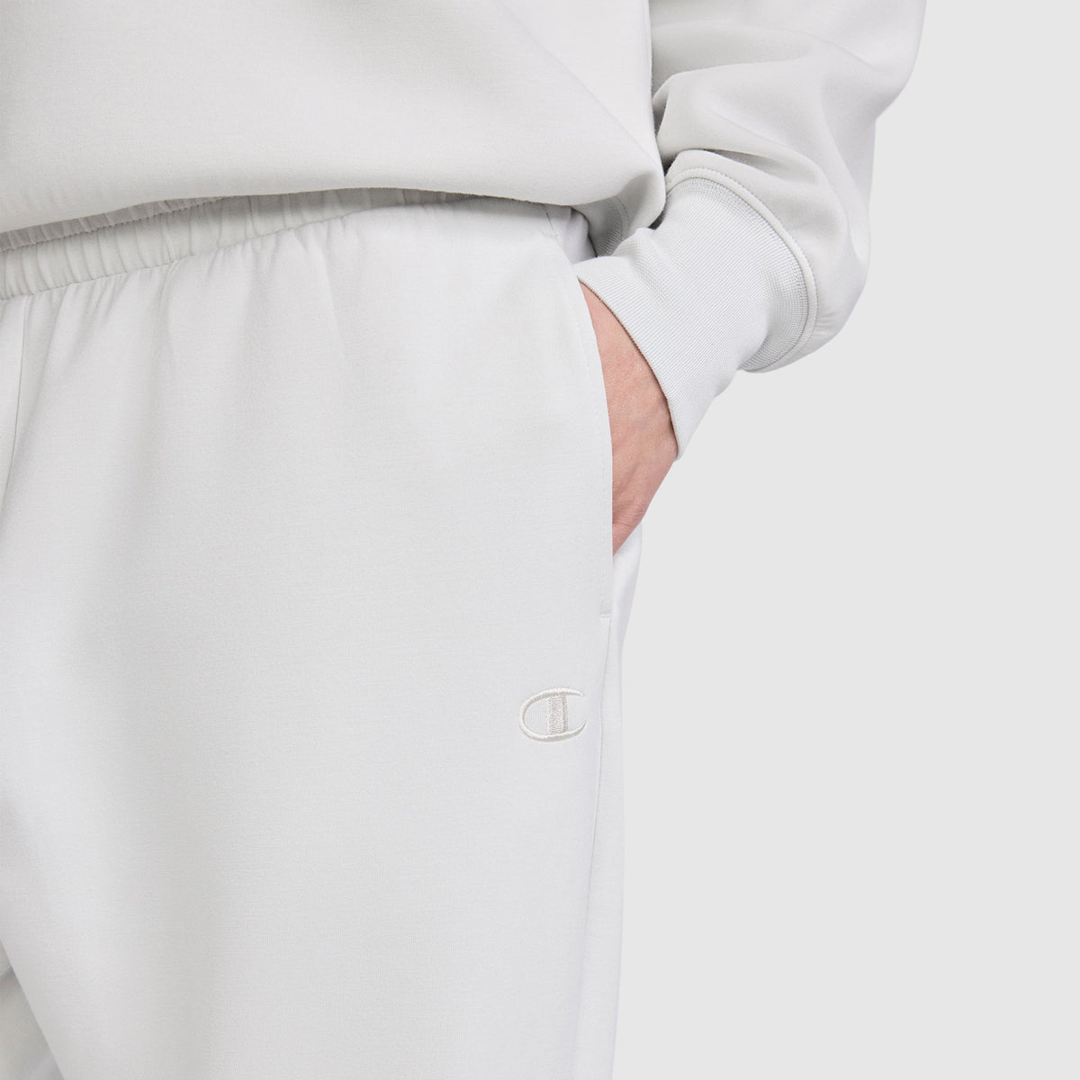 A person is wearing the Champion Taper Leg Jogger with Zippers in light gray, paired with a matching sweatshirt. Their right hand is in the pocket, and the Champion logo is visible on the joggers, which feature a tapered leg design. The background is plain and light-colored.