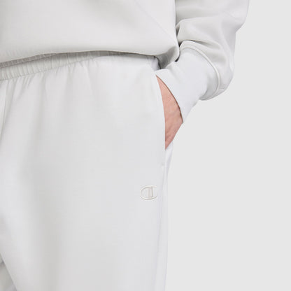 A person is wearing the Champion Taper Leg Jogger with Zippers in light gray, paired with a matching sweatshirt. Their right hand is in the pocket, and the Champion logo is visible on the joggers, which feature a tapered leg design. The background is plain and light-colored.