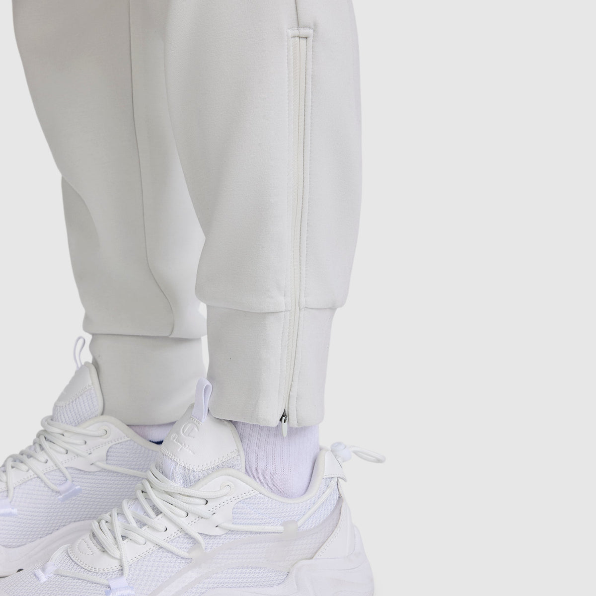 Close-up of a person wearing white sneakers, white socks, and the Champion Taper Leg Jogger with Zippers, featuring a tapered leg and side ankle zippers, standing on a plain light background.