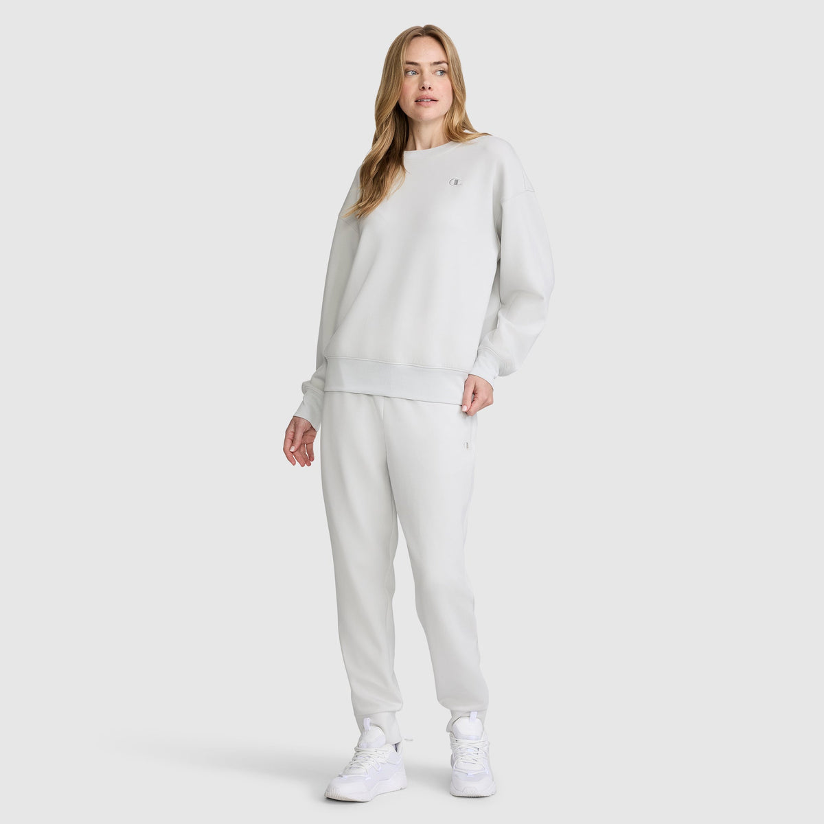 A woman stands against a plain white background wearing a Champion Taper Leg Jogger with Zippers and an oversized light gray sweatshirt, paired with white sneakers.
