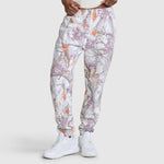 A model wears the Champion Powerblend Boyfriend Sweatpant with C Logo and an allover print featuring a gray, orange, and purple tree branch pattern reminiscent of camouflage. The jogger pants are styled with white sneakers and a white shirt, standing against a plain light gray background.