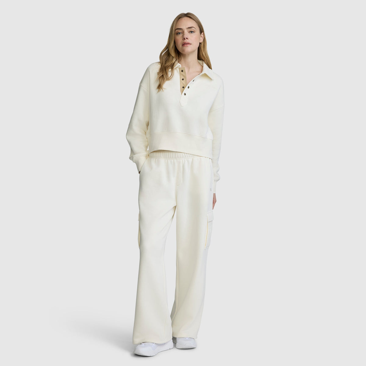 A person is wearing a cream-colored Champion Fleece Training Polo featuring long sleeves and buttons, styled with matching wide-leg pants. They have long, light brown hair and white sneakers, standing against a plain background.