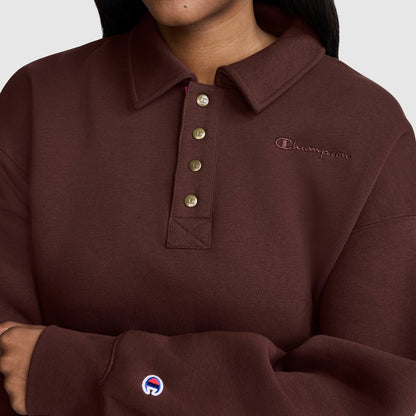 A person is seen wearing a Champion Fleece Training Polo in maroon, featuring a collared neckline and five-button placket. The stylish women’s fleece pullover has the Champion logo embroidered on both the left chest and sleeve. Only the lower face and upper body are visible.