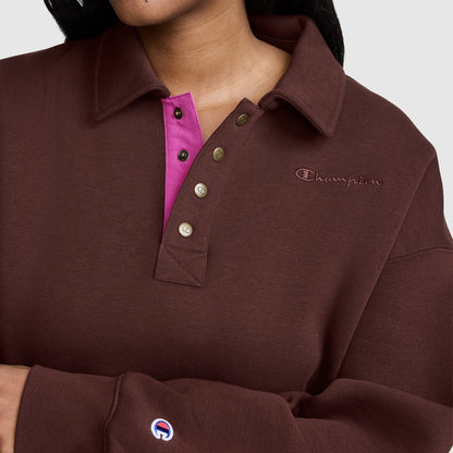 A person is wearing the Champion Fleece Training Polo in brown, featuring a collar, pink inner placket, and metallic buttons. Only the torso along with part of the face and arm are visible, highlighting this stylish women’s pullover that offers an athletic yet casual look.