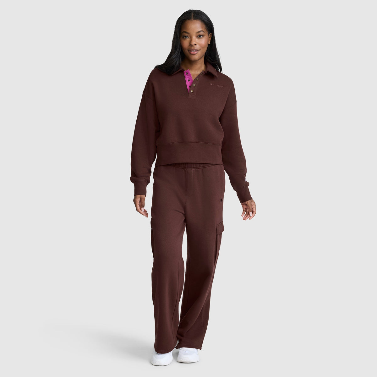 A woman stands against a plain light gray background, wearing the Champion Fleece Training Polo paired with matching pants and white sneakers.