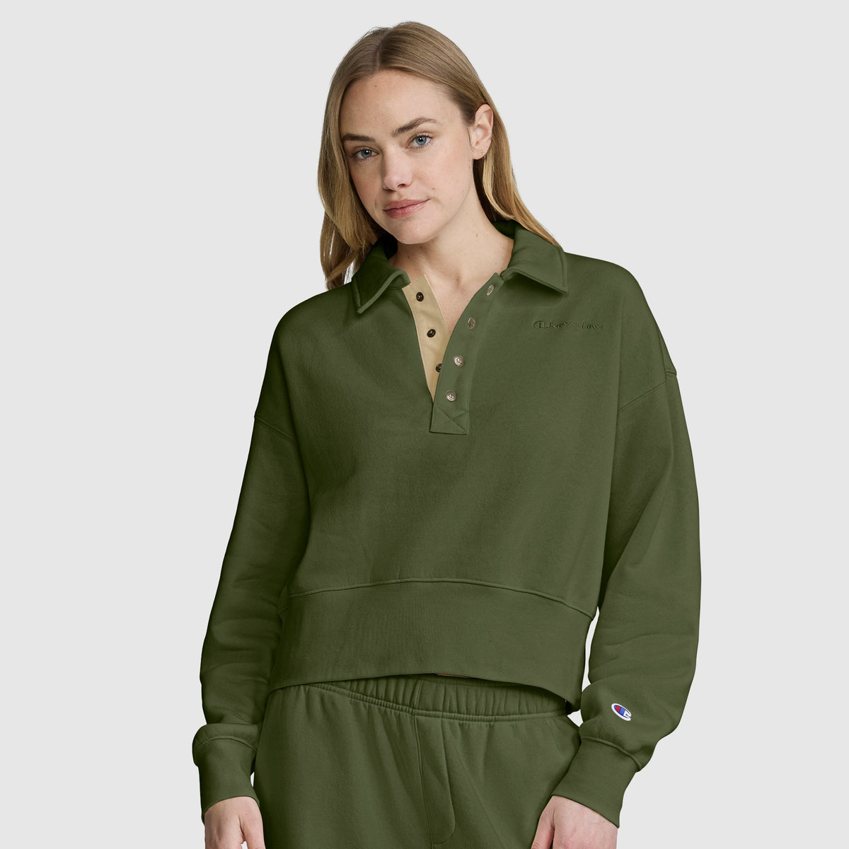 A woman with long blonde hair wears the Champion Fleece Training Polo in olive green, featuring a collar and matching pants, as she stands against a plain light gray background.