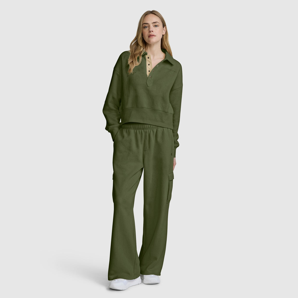 A woman stands facing forward, wearing a Champion Fleece Training Polo in olive green paired with matching wide-leg pants. She has long blonde hair and white sneakers, and is posing against a plain light gray background.