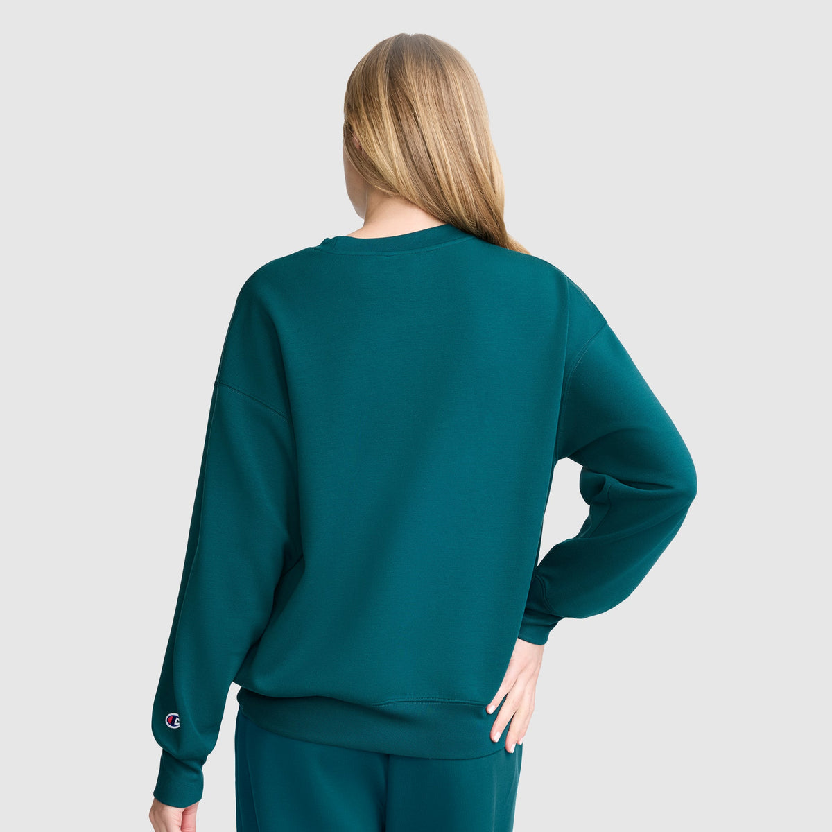A person with long blonde hair, seen from behind, wears a teal Champion Crewneck Pullover Sweatshirt and matching pants, creating an athletic silhouette. Their right hand rests on their hip against a light gray background.