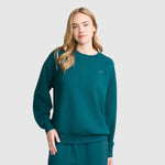 A woman with long blonde hair wears a teal Crewneck Pullover Sweatshirt and matching pants by Champion, standing against a light gray background and looking at the camera with a neutral expression. The athletic silhouette of the Champion Crewneck Pullover Sweatshirt adds a modern touch to her relaxed ensemble.